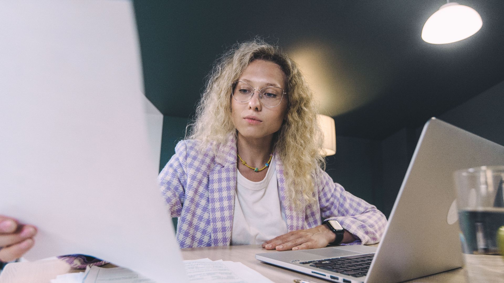 Woman in an office environment reviewing documents with focus, surrounded by technology.