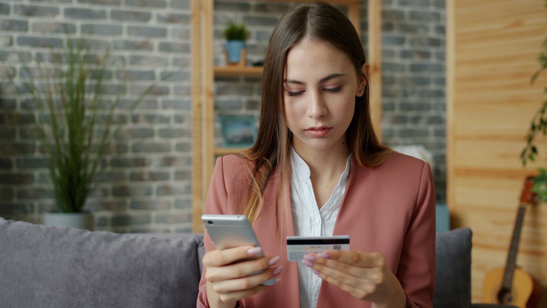 A young woman in a pink blazer shops online using a smartphone and credit card indoors.