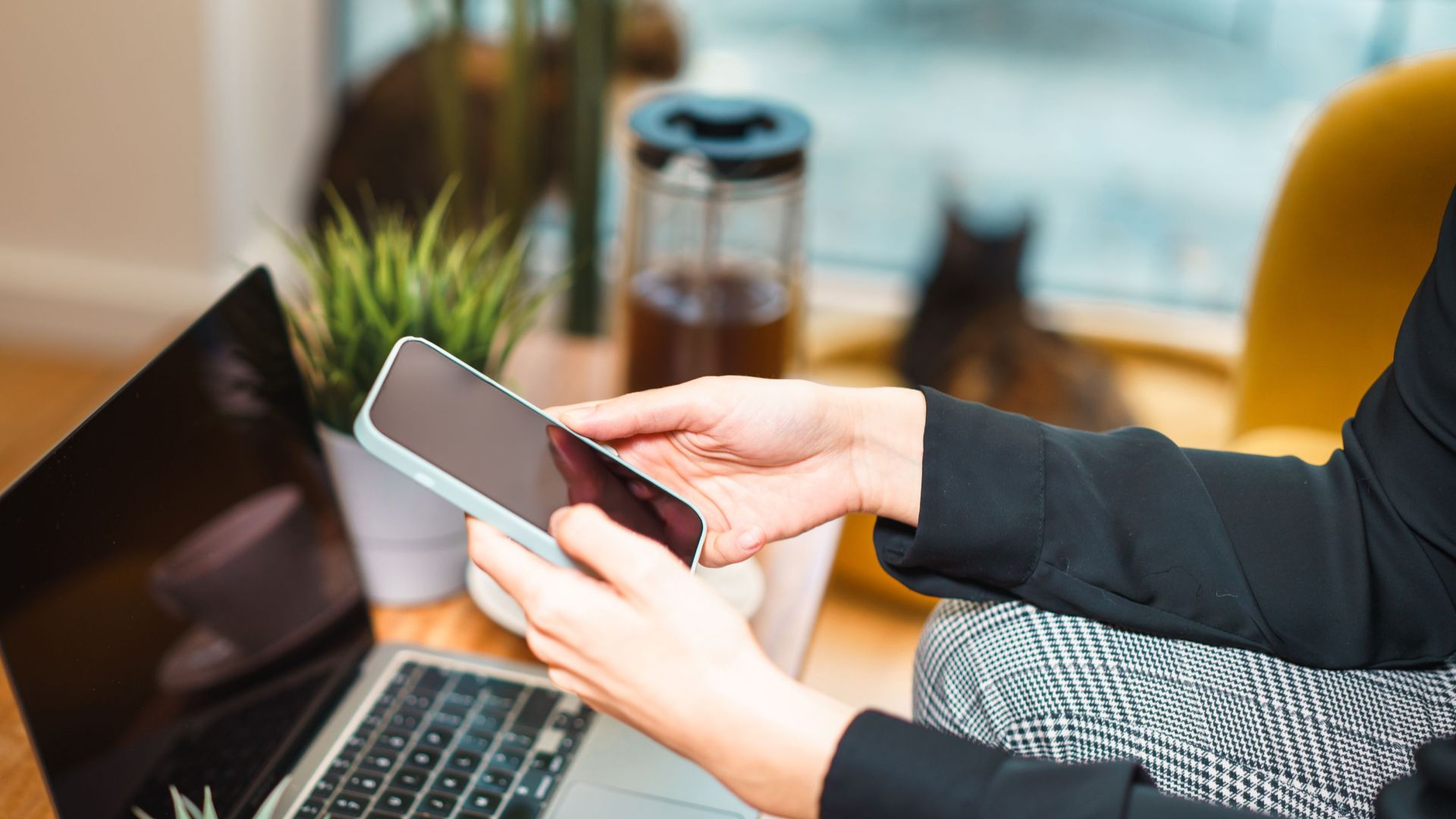Woman working remotely from home, using a smartphone and laptop at a cozy desk with a plant and cat in background.