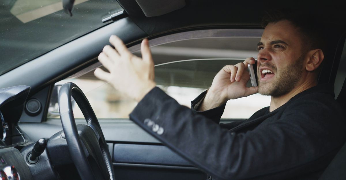 Stressed man gesturing while on phone in car, expressing frustration.