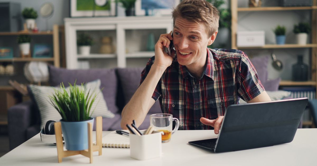 Man in a checkered shirt talks on phone in home office setting with laptop and coffee.