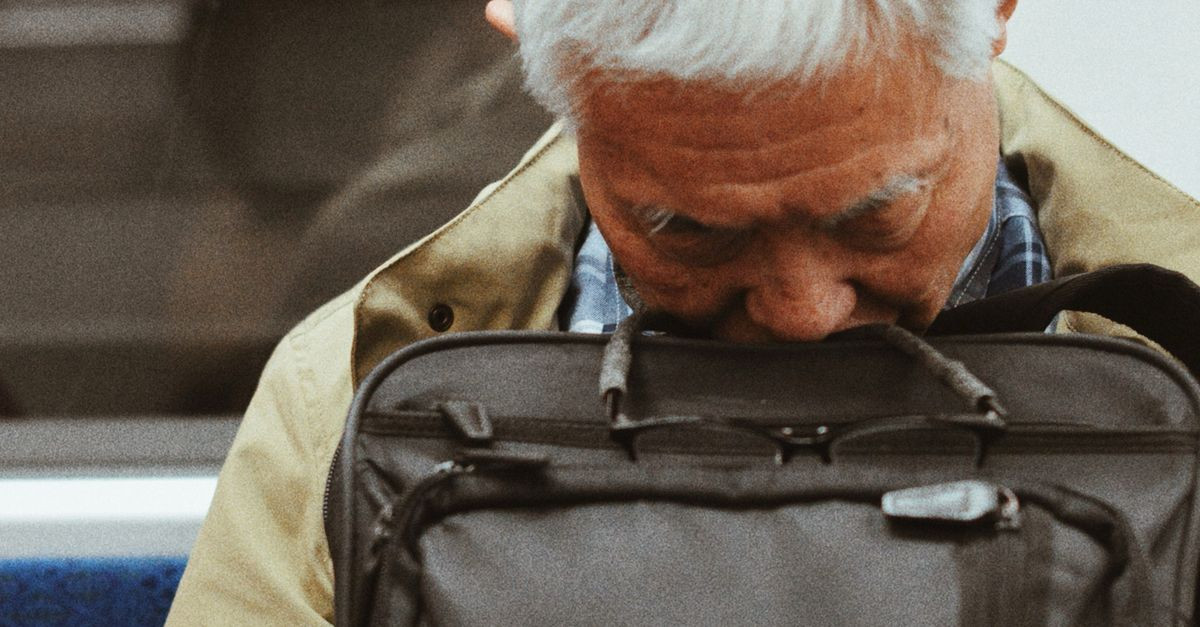 An elderly man asleep on a subway train, resting his head on a backpack.