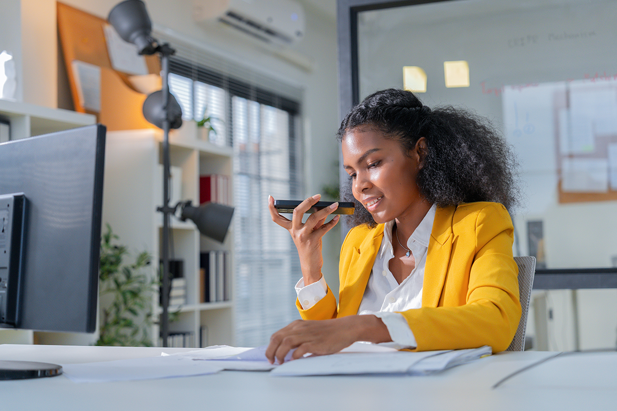 Confident professional businesswoman using smartphone voice message while reviewing documents at modern office desk. Young corporate employee working with technology, communication tools