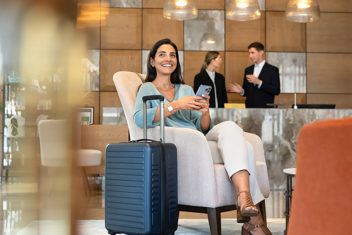 Smiling female hotel guest sitting in reception using smartphone to check messages, confirm reservation, or complete online check-in. Digital services, modern travel app usage, mobile communication