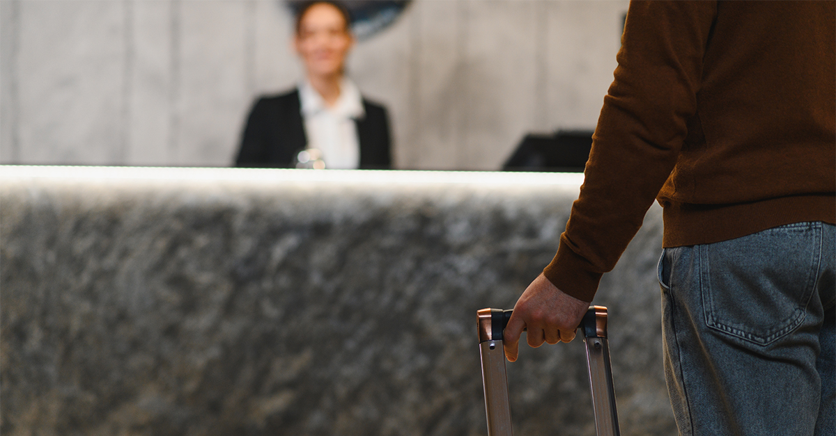 Traveler standing at a hotel reception desk, holding luggage, communicating with the blurred receptionist