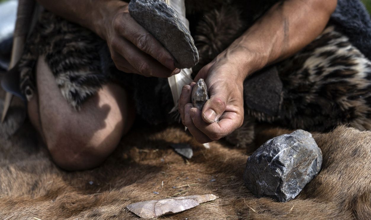 Getty Images - 1160507309 - Neanderthal Cave man works on a flint ax in a wood.