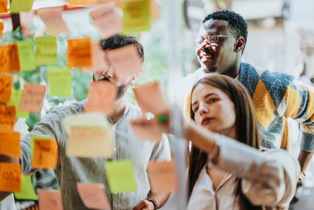 Creative professionals work on a project, utilizing a glass board and sticky notes. Successful teamwork and communication lead to a modern, persistent corporation.