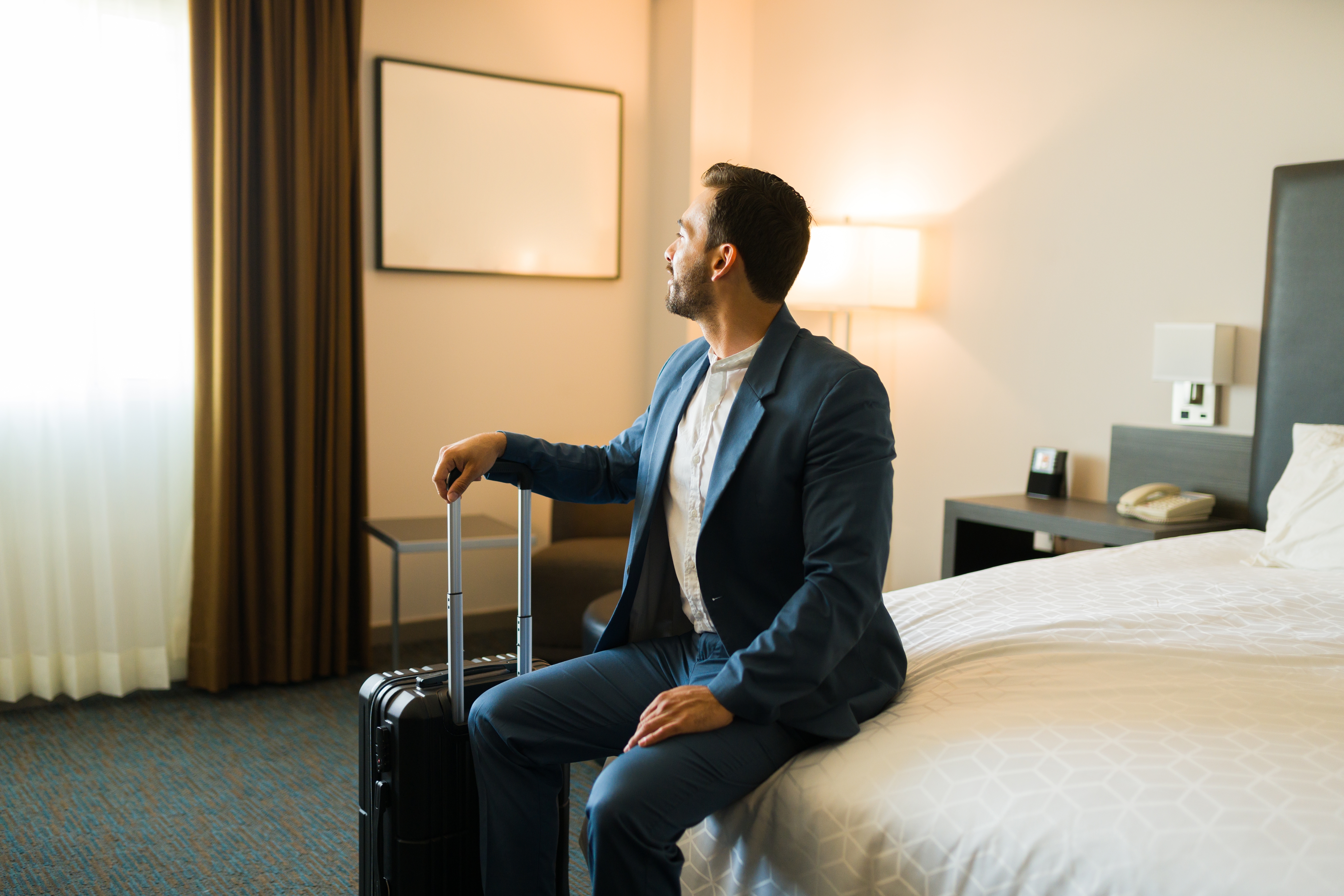 Handsome young businessman in hotel room sitting on bed with suitcase and looking out the window