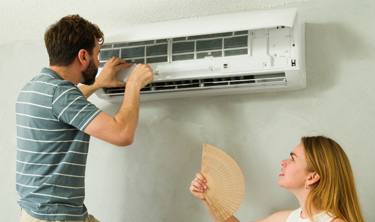 Caucasian man standing on a ladder installing or repairing a mini split ac unit while a woman fans herself, waiting for cool air