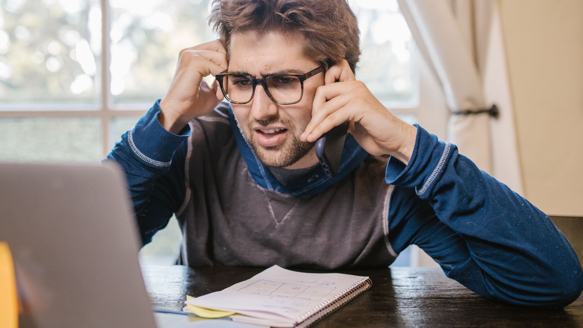 Man in glasses using laptop and smartphone at home, appearing confused.