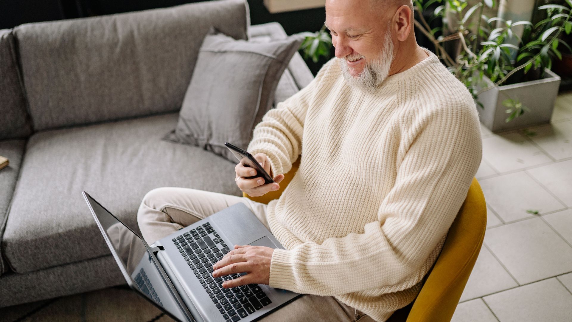 A smiling elderly man sits comfortably indoors, using both a phone and laptop, with a cozy ambiance.