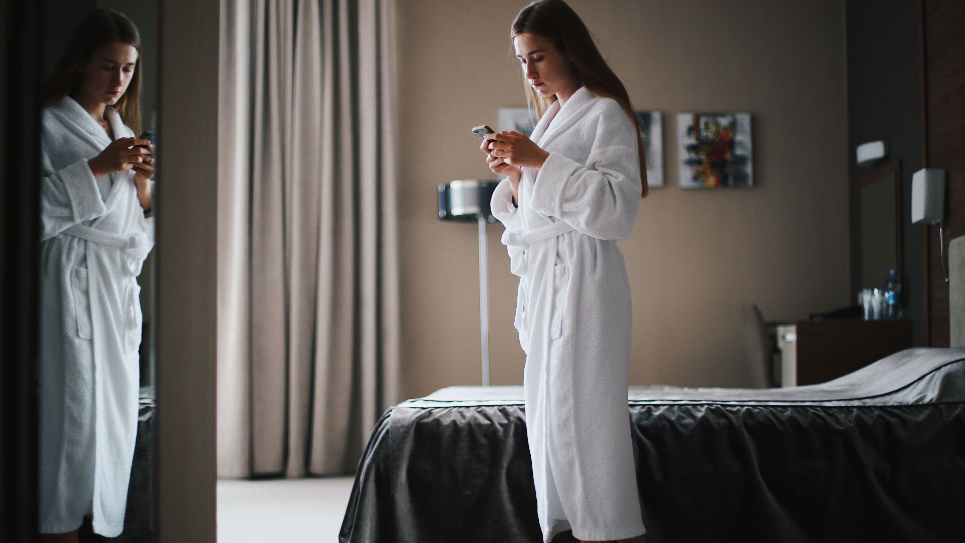 Woman in bathrobe looking at smartphone in a modern hotel room, near a mirror.