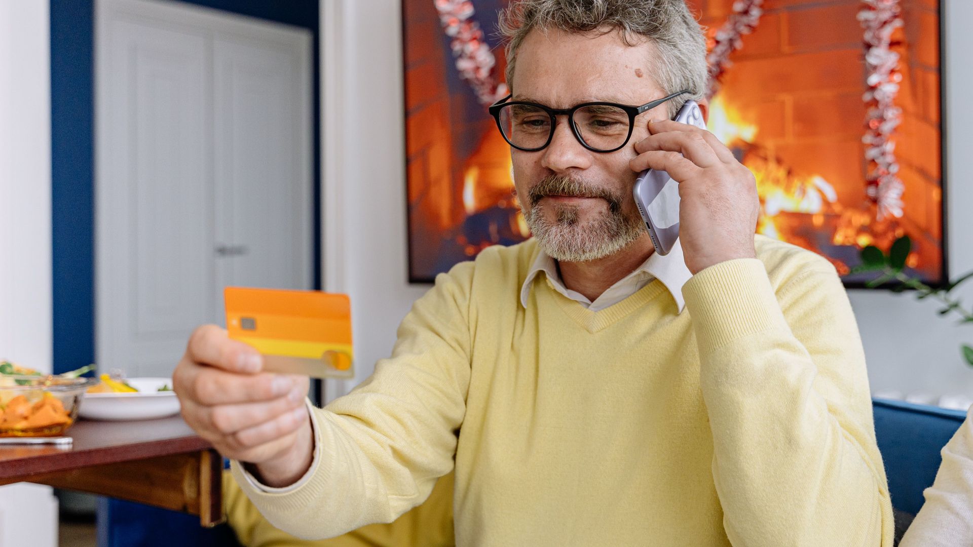 Elderly man using phone while holding a credit card indoors near a cozy fireplace.