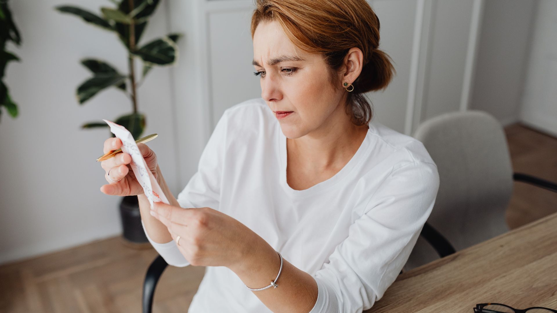 A concerned adult woman examines a receipt while sitting at her office desk, indicating financial scrutiny.