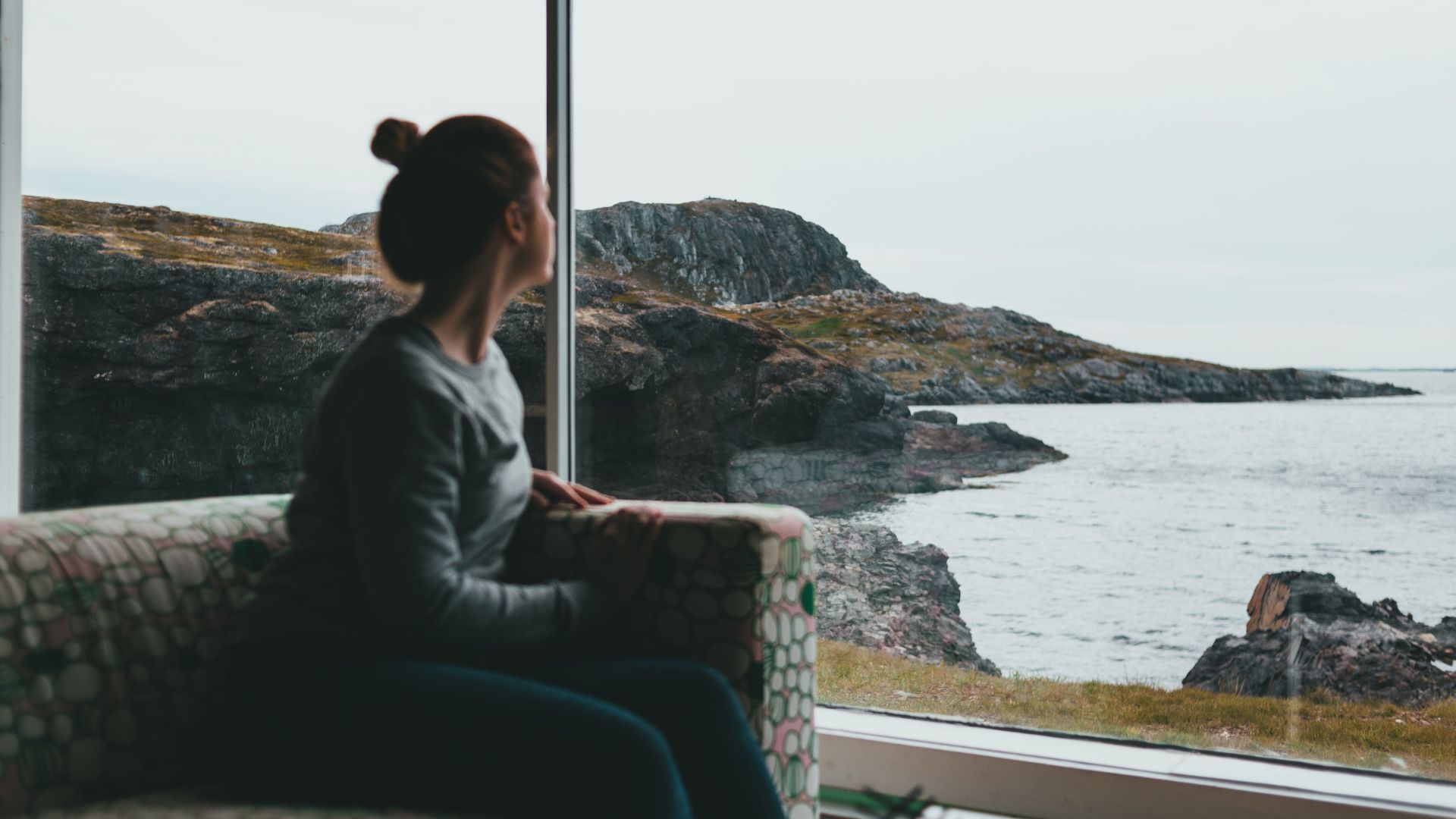 A woman sits indoors looking through a window at a rocky coastal landscape.