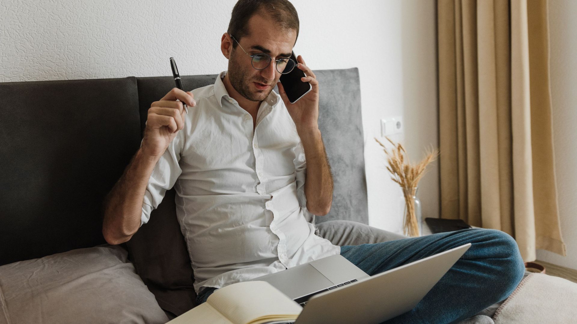 Adult male multitasking at home office, using laptop and smartphone while sitting on bed.
