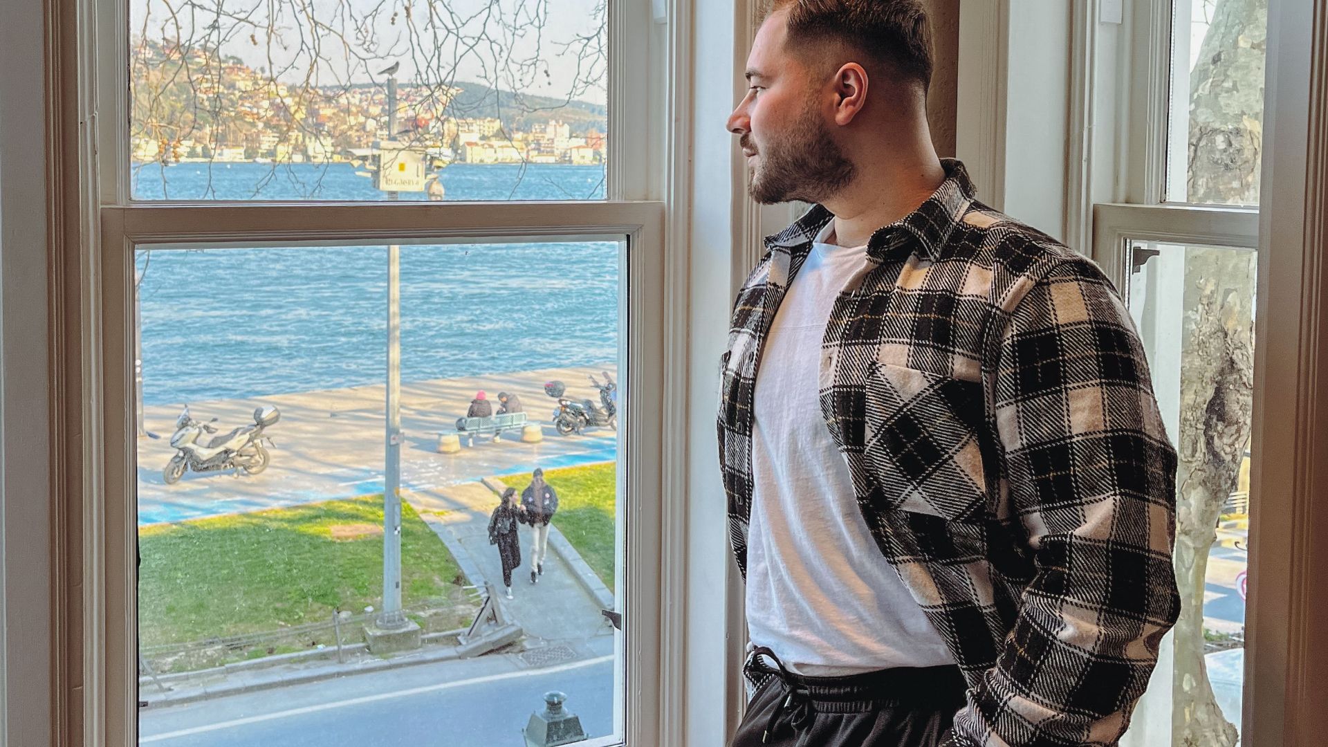 A man in casual wear gazes out a window, overlooking a scenic waterfront on a sunny day.