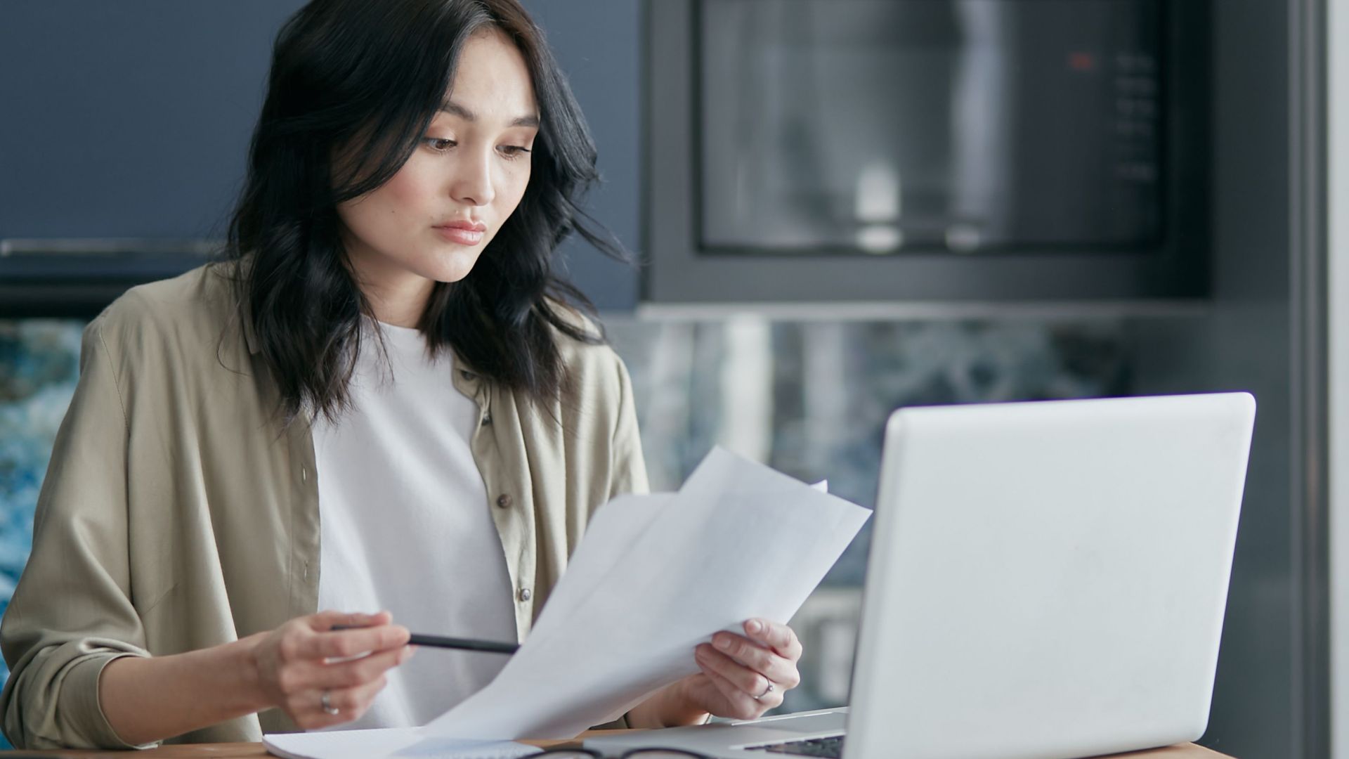 Concentrated woman reviewing documents and working on a laptop at home office setup in kitchen.