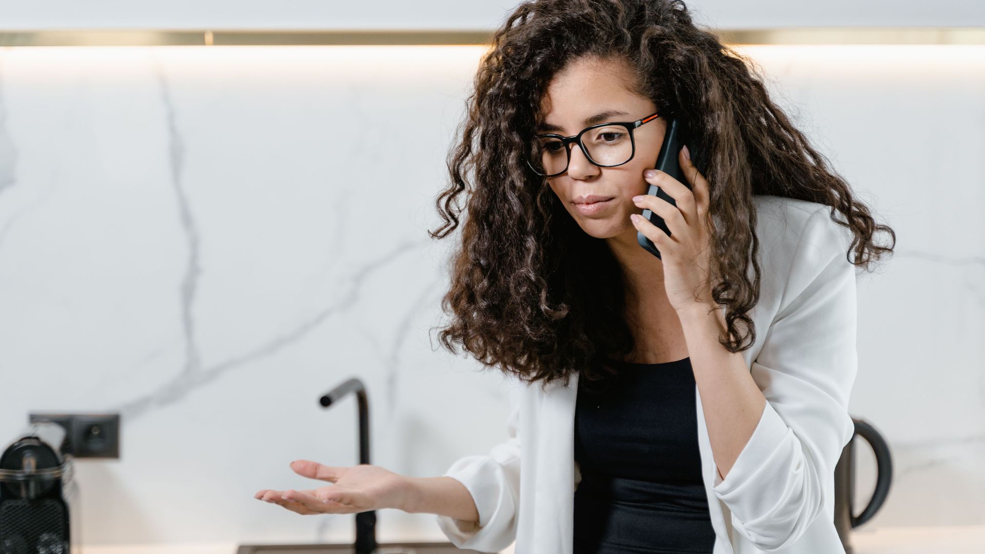 A woman with curly hair and glasses makes a phone call in a sleek, modern kitchen setting.