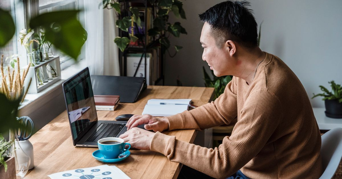Man in brown sweater working on a laptop with coffee on a wooden desk in a home office setting.