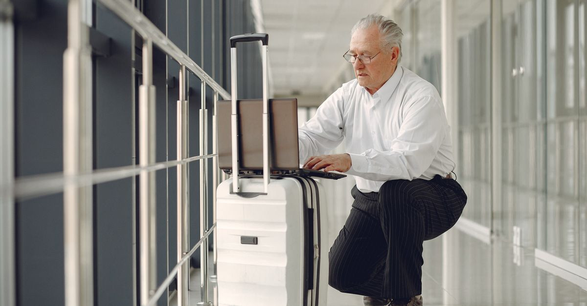 Full length of focused mature male in formal wear using laptop on baggage while having semisitting position in airport corridor