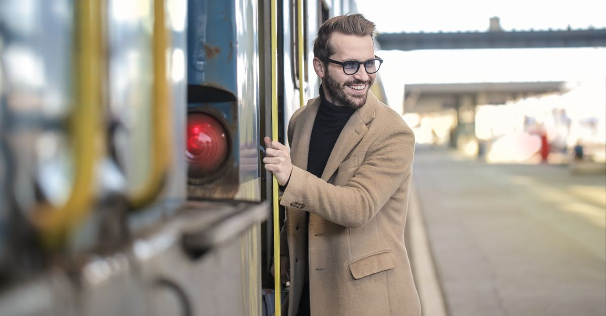 Man in a coat with glasses boards a train at an outdoor station, smiling.