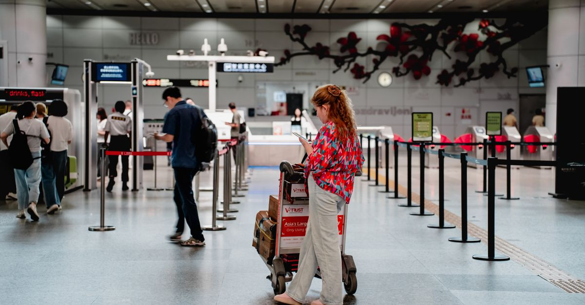 A traveler checks her phone while waiting with a luggage cart at a busy airport terminal in Guangzhou.