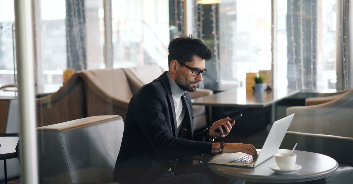 A businessman multitasking with laptop and phone in a stylish café setting.