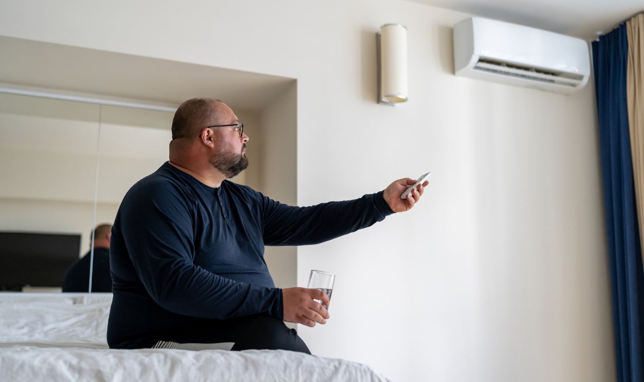 Frustrated overweight man suffering from heat hold glass of water includes conditioning at home. Unwell fat male sweats while sits on bed feel blood pressure try cooling in hotel room. Hot air indoor