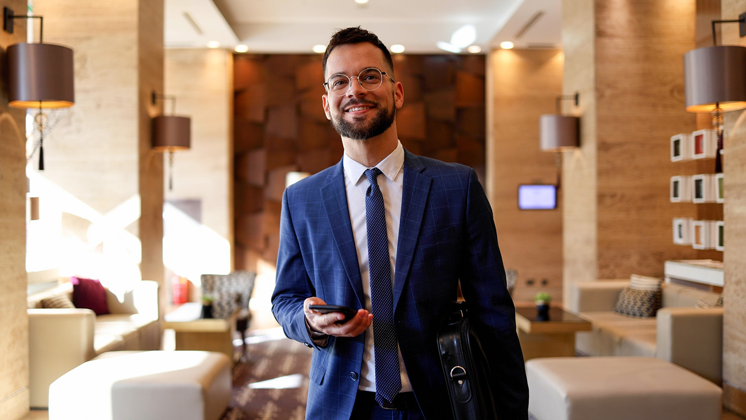 Smiling businessman in suit holding phone and brief case, walking through hotel lobby, looking at camera confidently