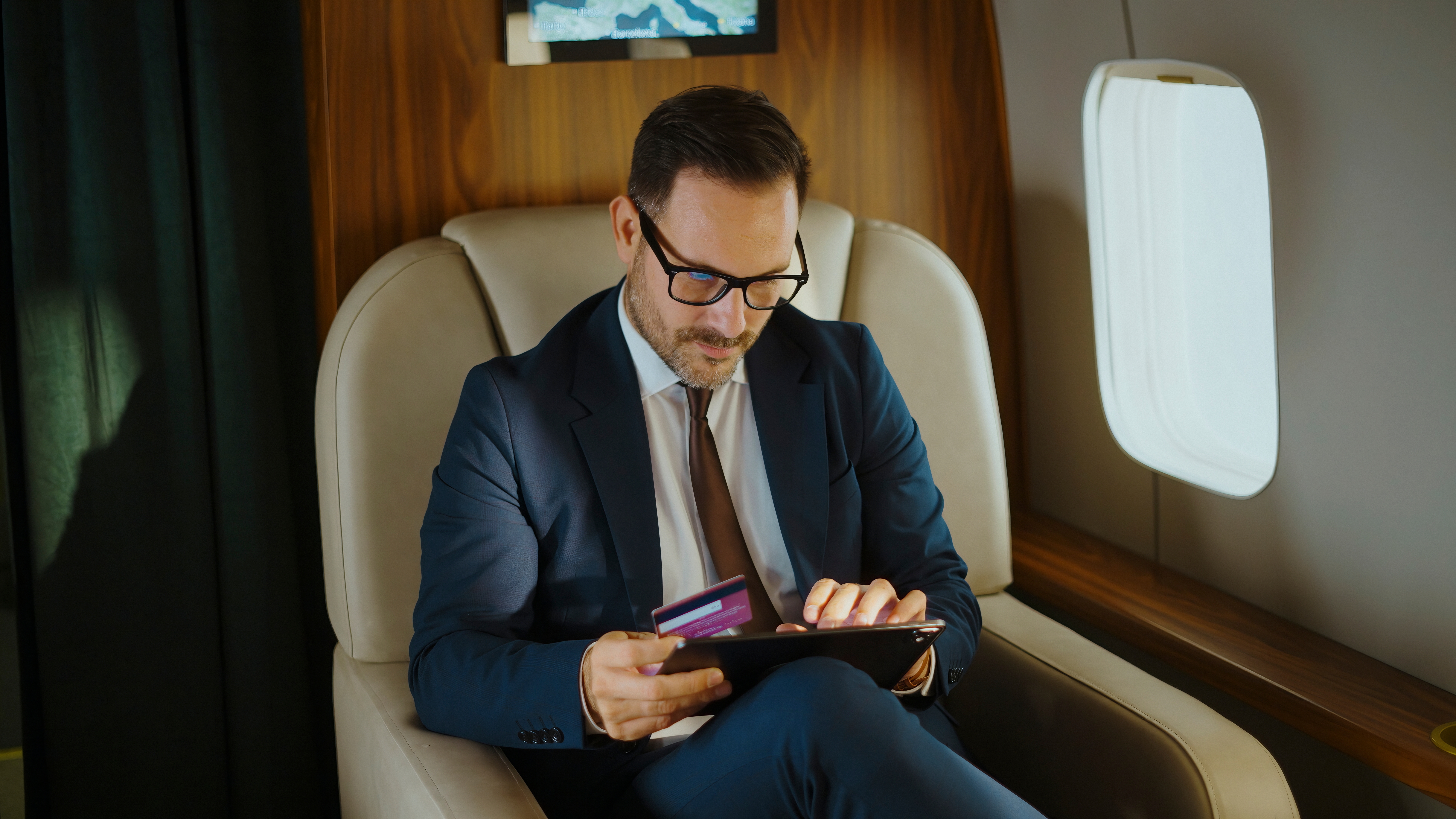Business executive in suit and glasses using a tablet and credit card to shop online while traveling in the luxurious cabin of a private jet, showcasing modern mobile commerce