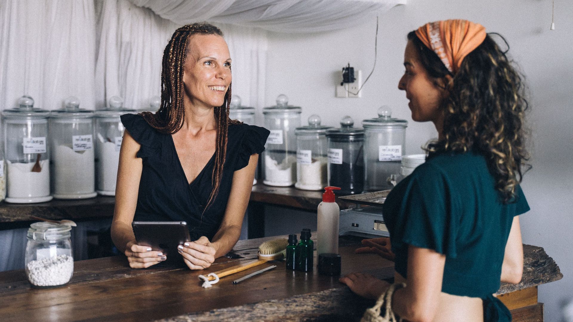 Woman using a tablet to assist customer in eco-friendly store with jars and eco products.