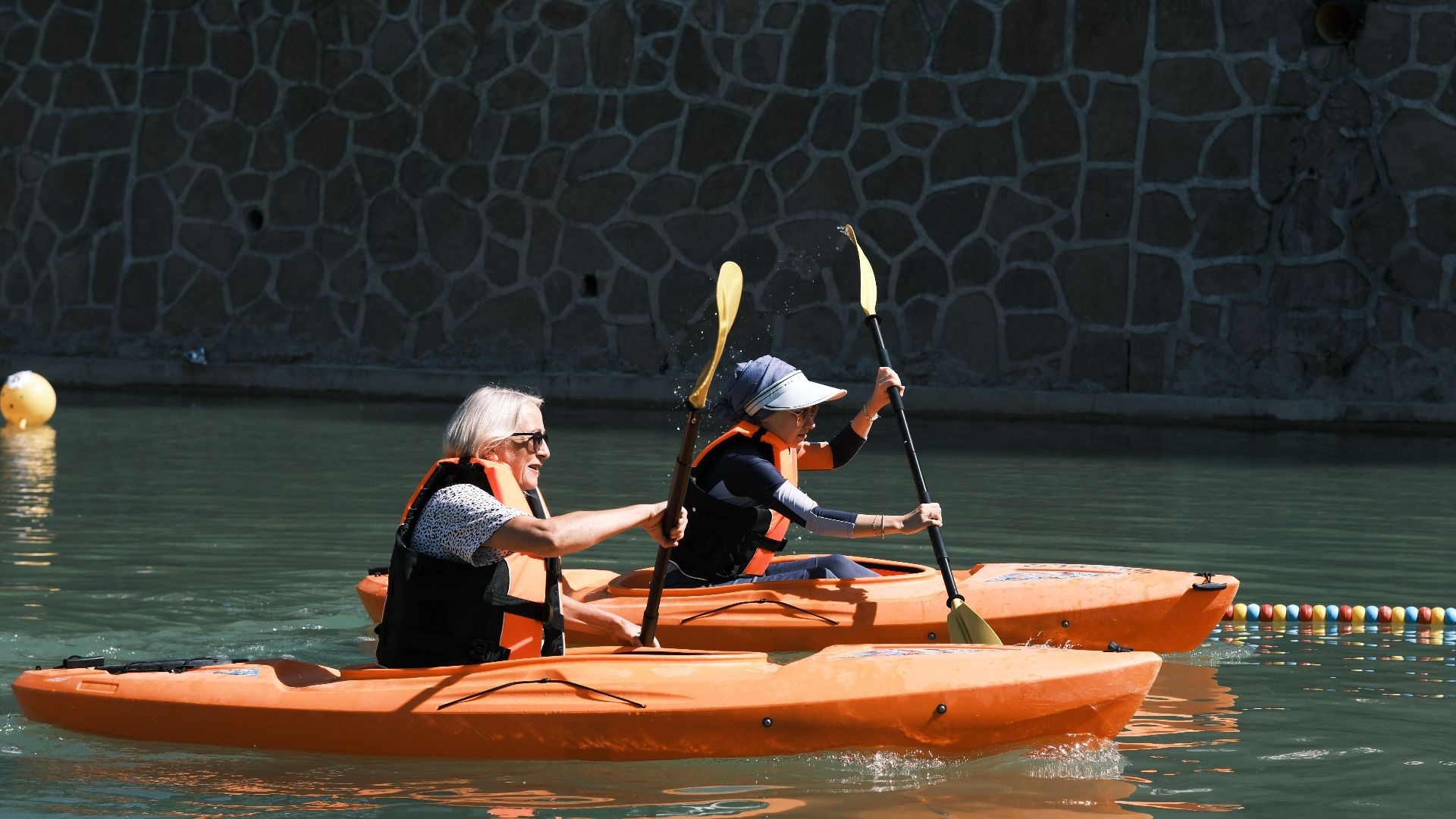 Two people kayaking in orange boats on a sunny day by stone wall.