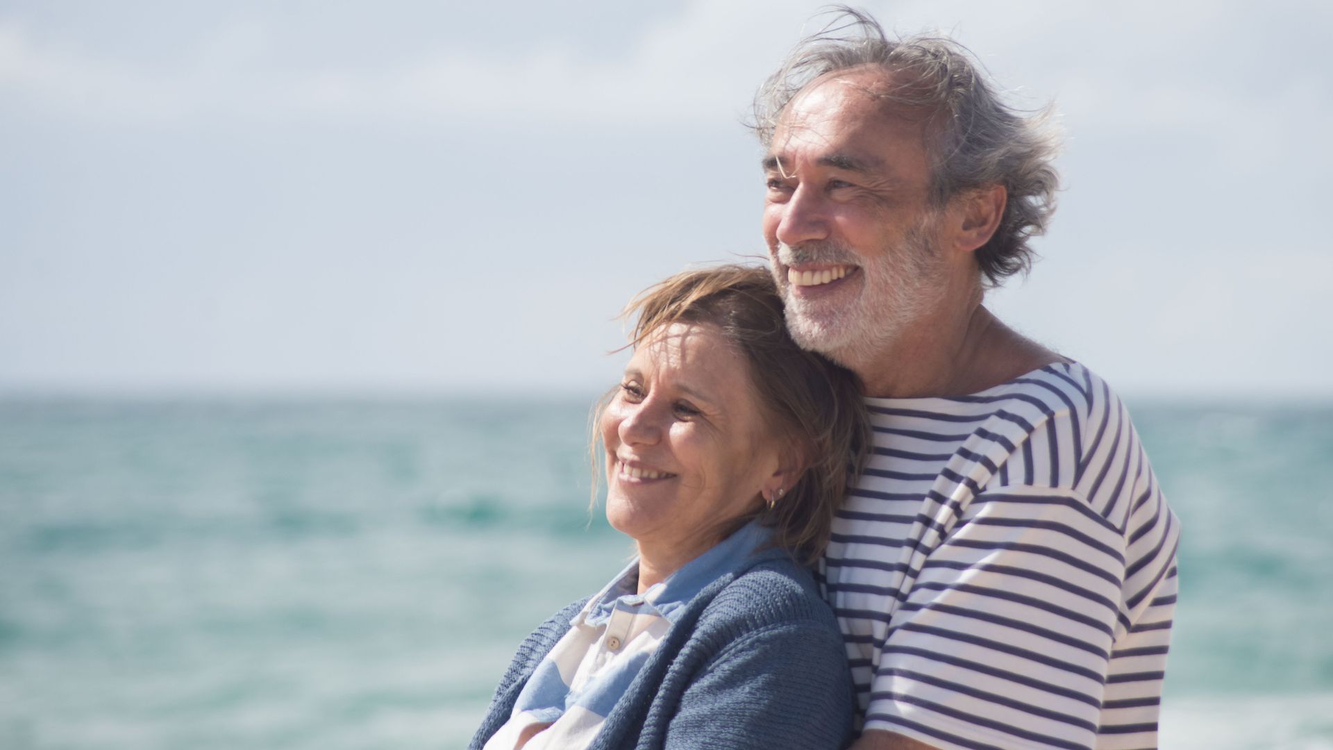 Happy senior couple embracing each other by the ocean, enjoying a sunny day in Portugal.