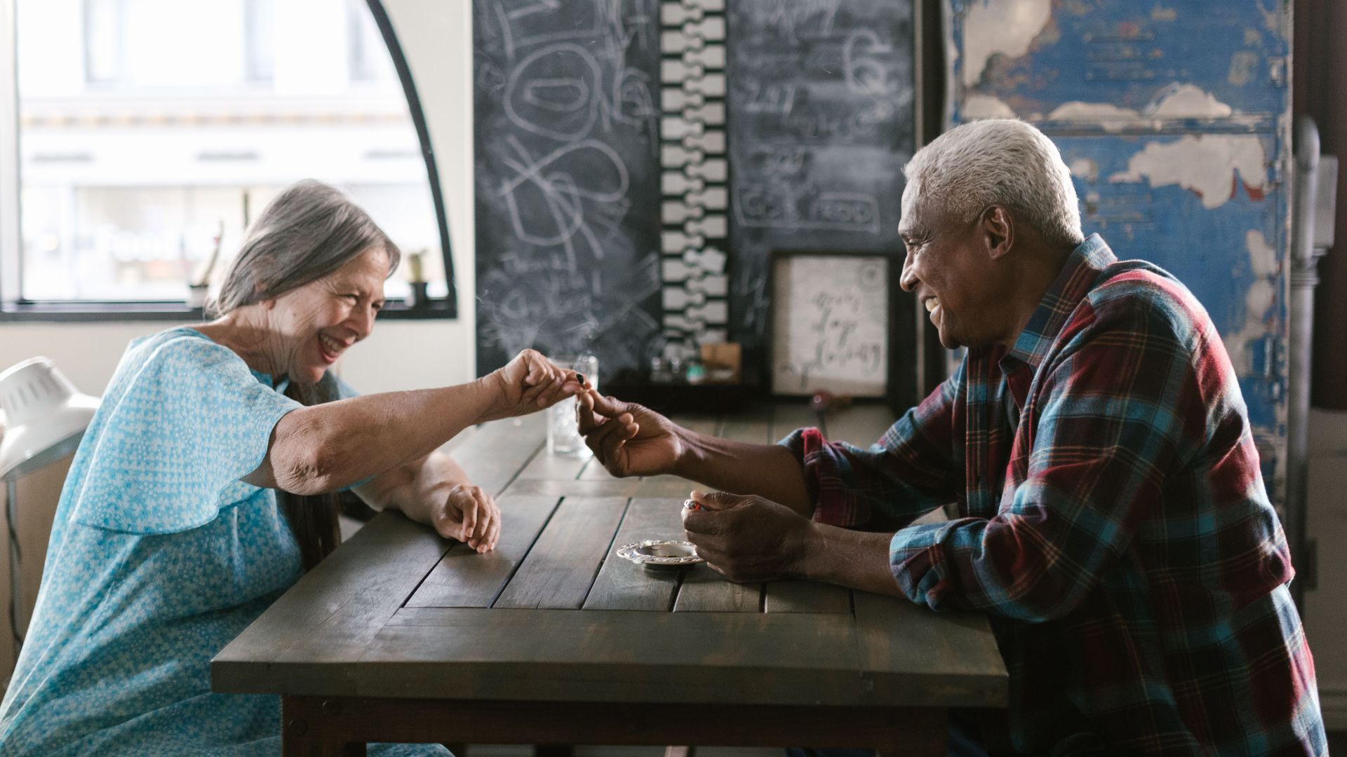 Elderly couple laughing and playing a game at a table indoors, enjoying each other's company.