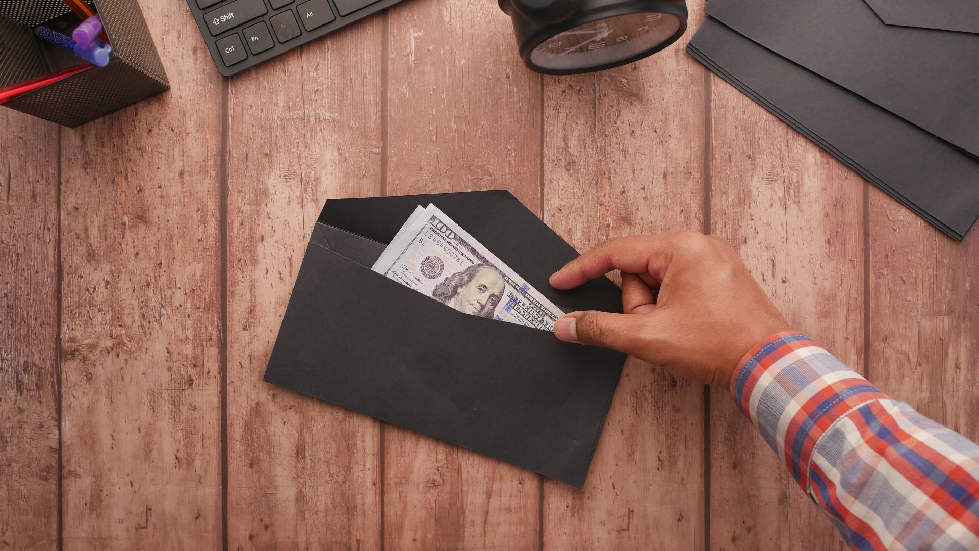 A hand placing a 100 dollar bill in a black envelope on a wooden desk with stationery, keyboard, and alarm clock.