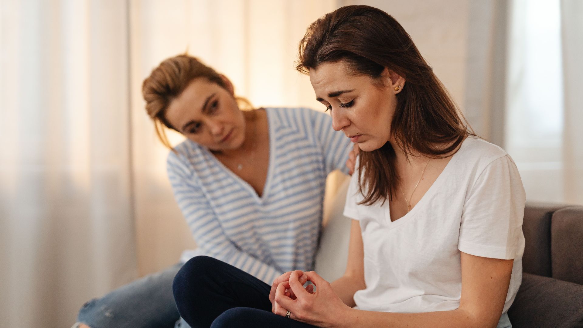 Two women sitting on a couch, sharing emotional support and connection in a warm living room setting.