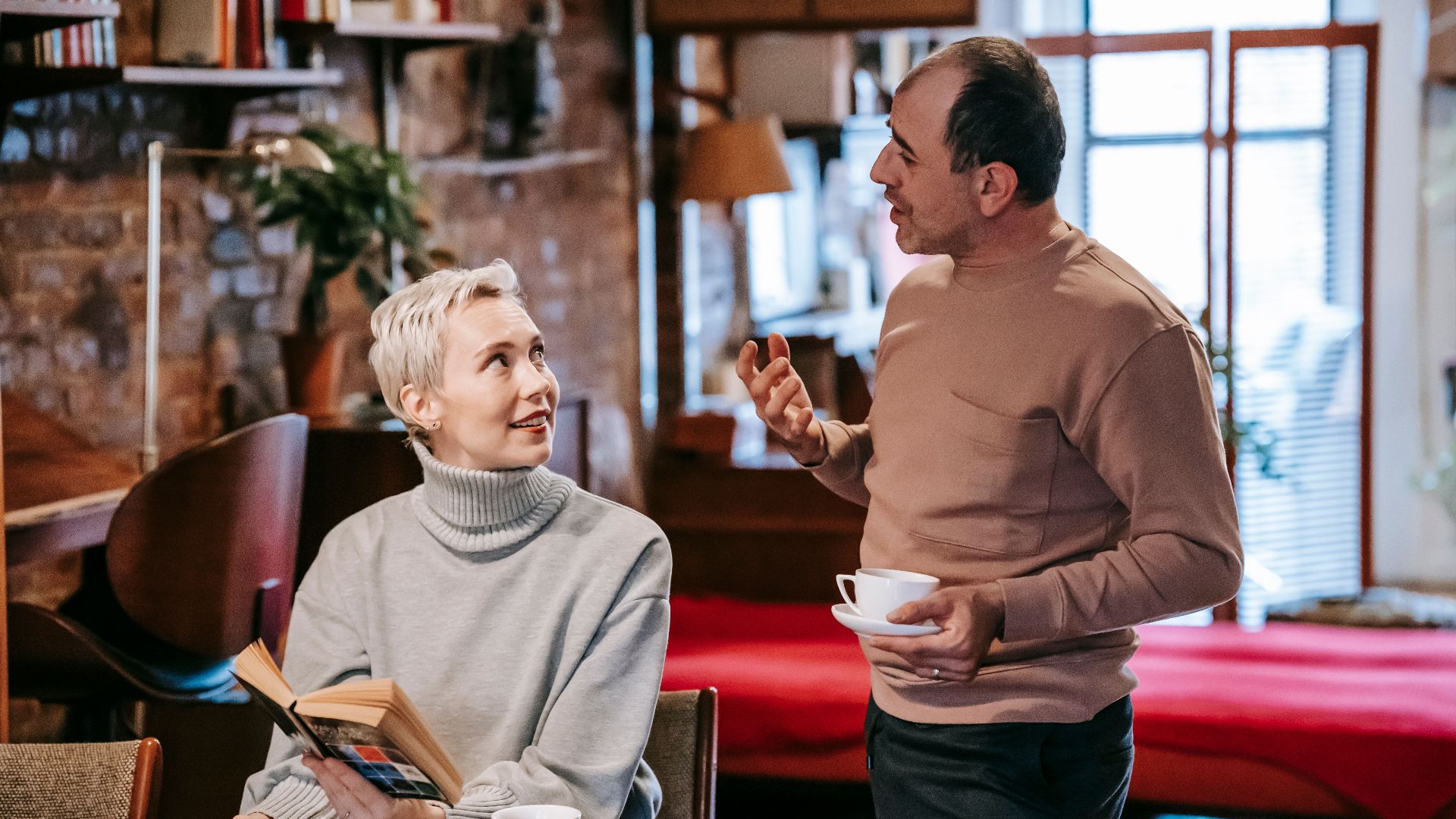 Positive diverse middle aged spouses in casual clothes drinking coffee and discussing interesting book while spending weekend together at home