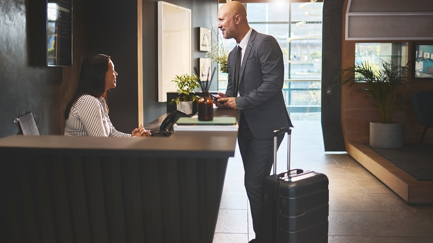 Receptionist, people and man in hotel with check in for accommodation on business trip. Woman, employee and male client or customer with smile in help desk for booking and hospitality as guest