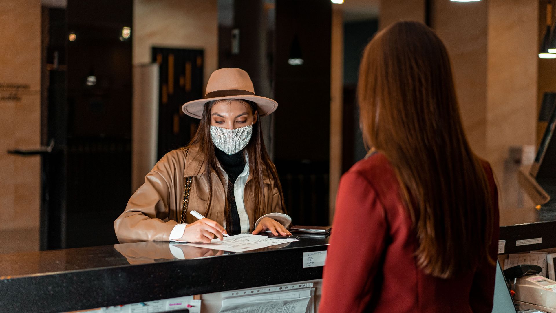 A woman in a hat and face mask checks in at a hotel reception, interacting with a receptionist.