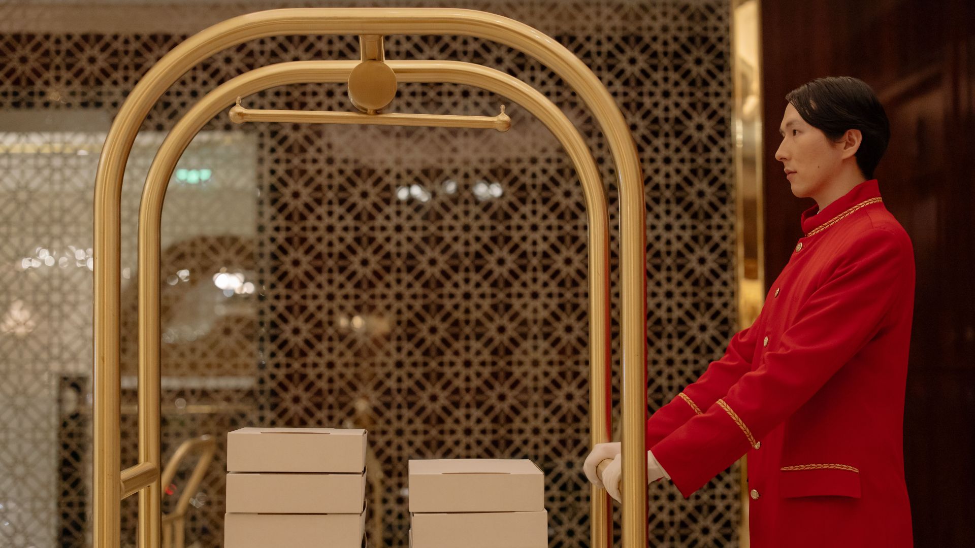 A hotel bellboy in uniform pushes a luggage cart with boxes indoors, demonstrating hospitality service.