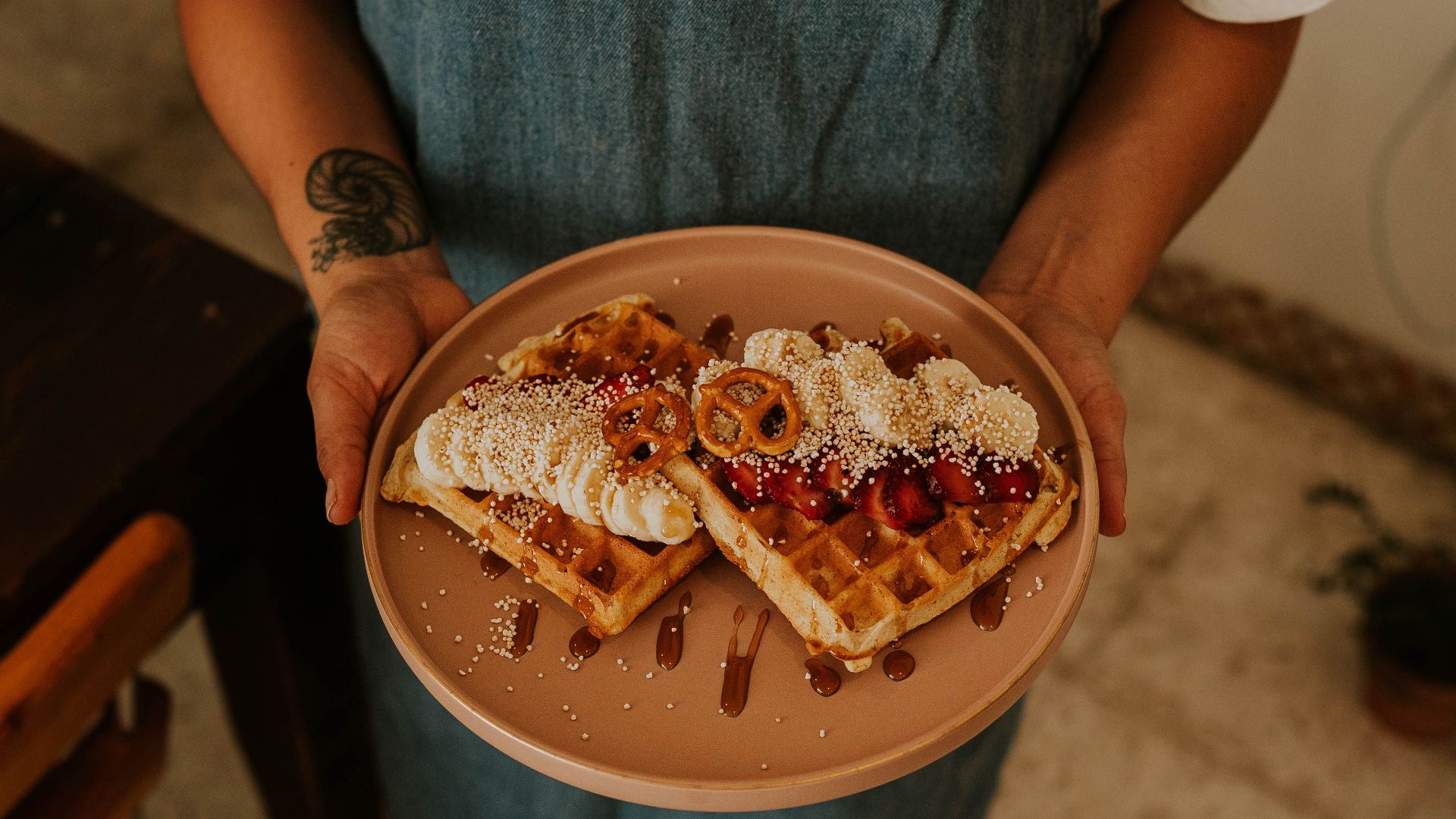Close-up of a person holding a plate of waffles topped with cream, berries, and pretzels in a cozy kitchen setting.
