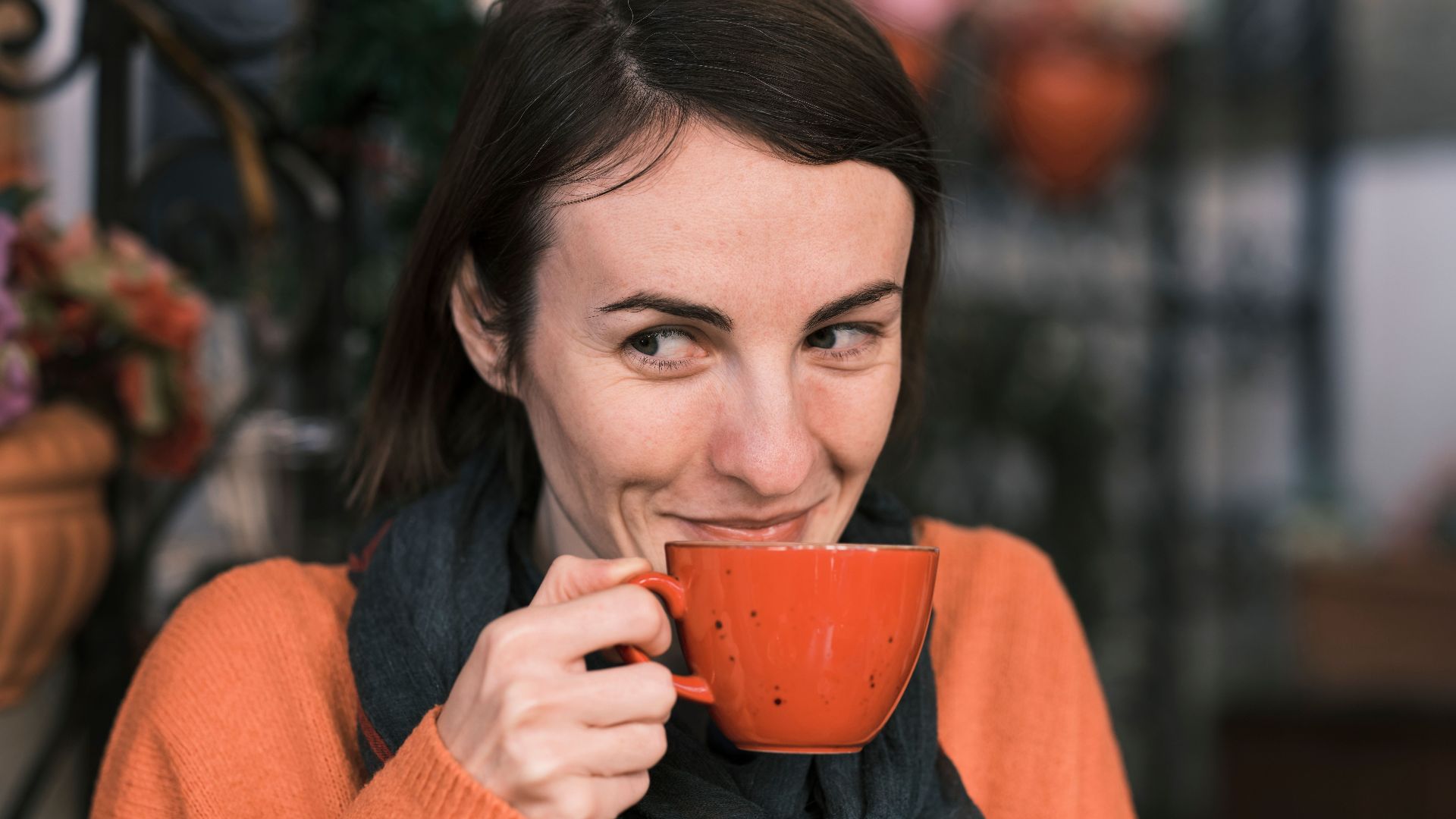 Cheerful woman in orange sweater sipping coffee outdoors, exuding warmth and happiness.