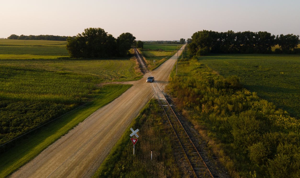 A quiet rural intersection with railroad tracks and a lone car, surrounded by lush farmland near Minneapolis, Minnesota