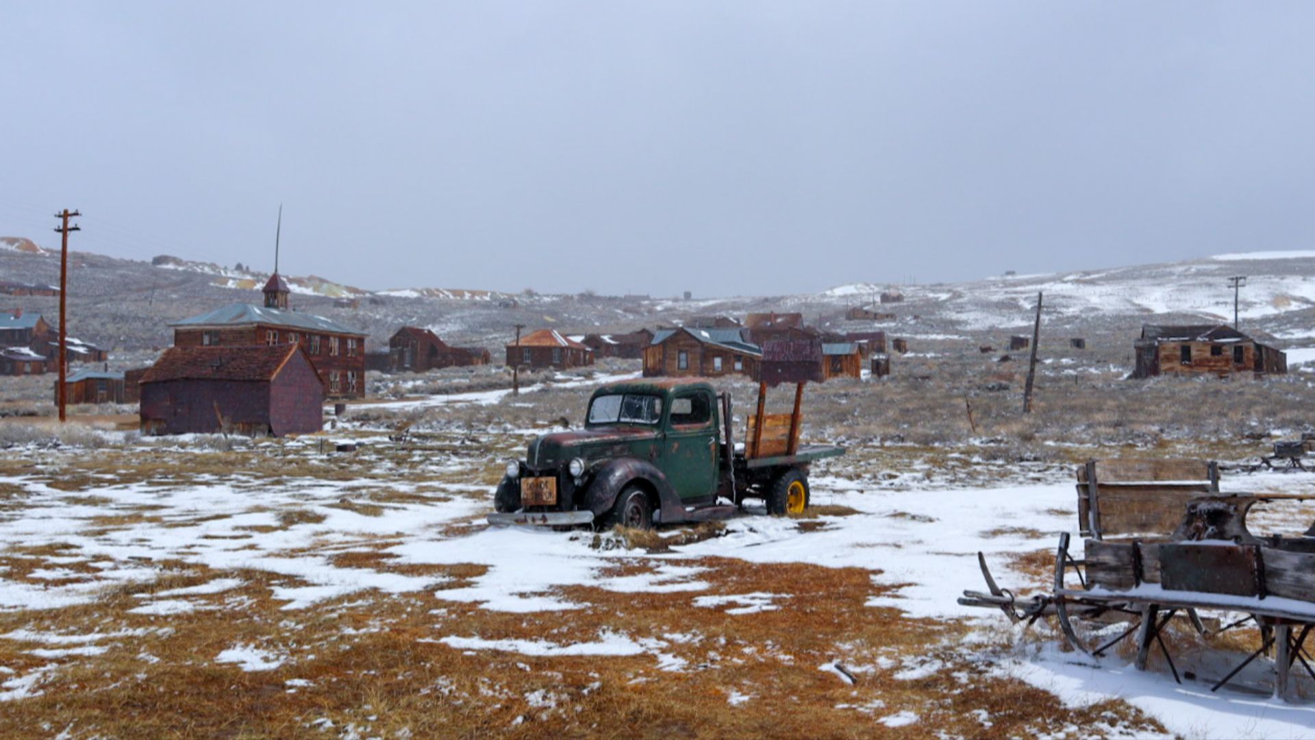 An old rustic green truck in the middle of the frame sits on snowy ground. Old wooden buildings are spread throughout the background on snowy hills beneath a plain gray sky.