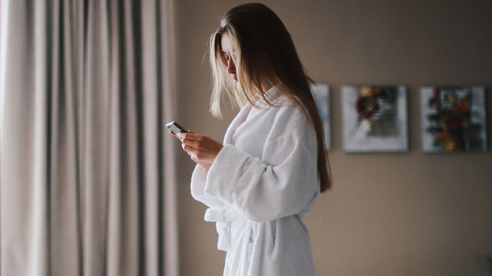 Woman standing in a hotel room wearing a bathrobe, focused on her smartphone for communication or relaxation.