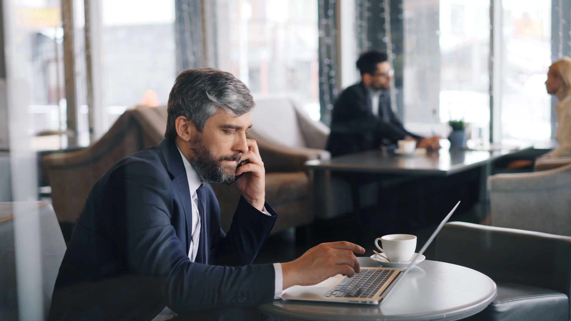 Businessman on phone and laptop in a modern cafe setting, focused and multitasking.
