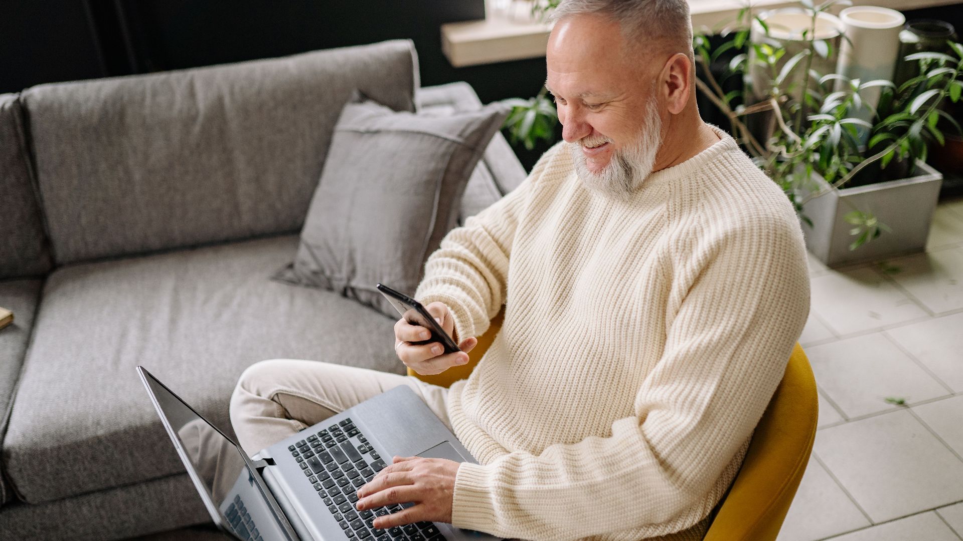 A smiling elderly man sits comfortably indoors, using both a phone and laptop, with a cozy ambiance.