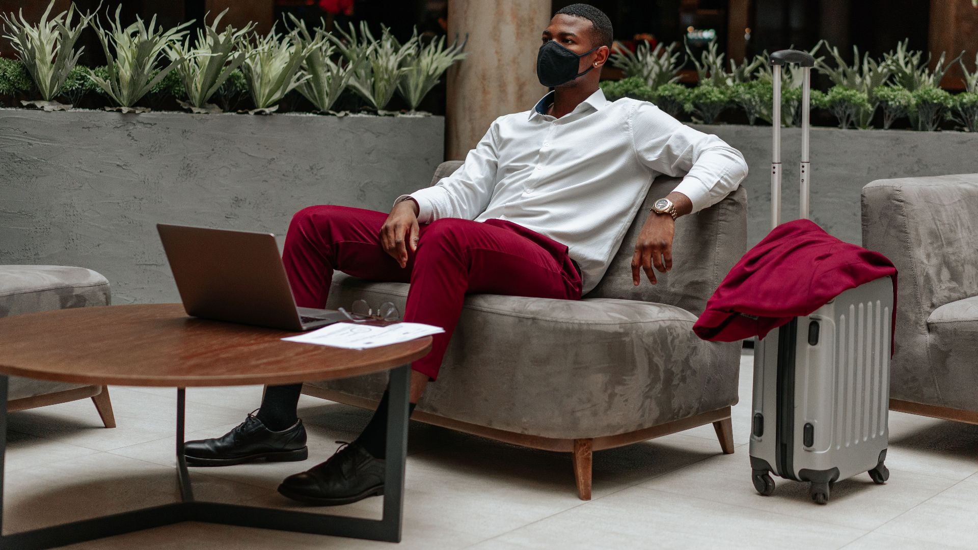 Man wearing facemask, sitting in hotel lobby with laptop and suitcase, adapting to new normal.