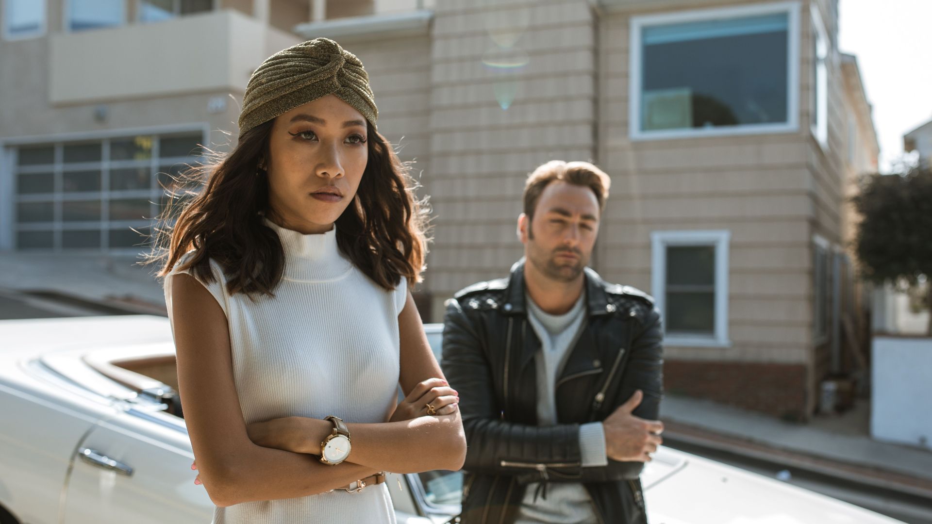 A couple appears upset, standing apart on a sunny urban street near a vintage car.